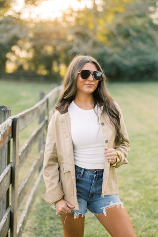 Woman standing outdoors in a field wearing sunglasses, beige jacket, white top, and denim shorts.