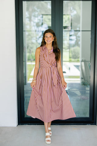 A woman stands in front of a glass door wearing a sleeveless midi dress with vertical red, pink, and white stripes, paired with white slide sandals.