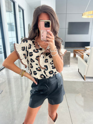 Woman taking a mirror selfie wearing a leopard print top and black shorts in a modern living room.