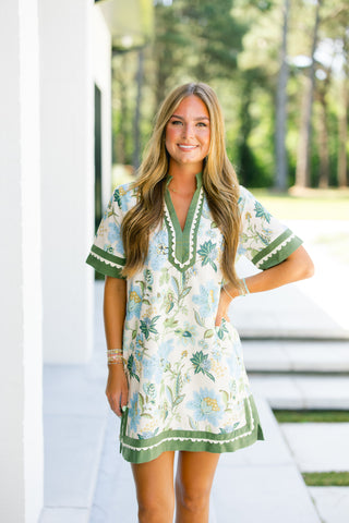Woman wearing a floral dress with green, sky blue and ivory accents standing outdoors.