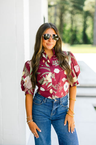 Woman wearing a burgundy floral blouse and blue jeans with sunglasses outdoors.