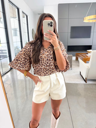 Person taking a mirror selfie wearing a leopard print top and white shorts in a modern living room.