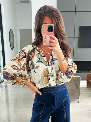 Person wearing a floral blouse and blue jeans taking a mirror selfie in a modern living room.
