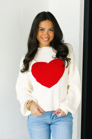Woman wearing a white sweater with a red heart design against a white wall.
