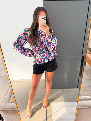 Person wearing a colorful floral top and black shorts taking a mirror selfie.