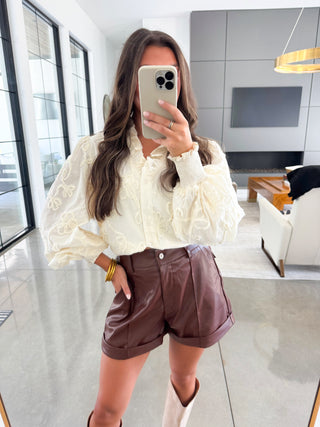 Woman taking a mirror selfie wearing a white blouse and brown shorts in a modern living room.