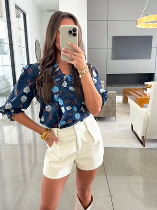 Woman taking a mirror selfie wearing a blue floral blouse and white shorts in a modern living room.