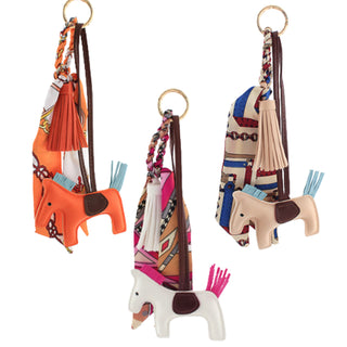 Horse-shaped bag charms with braided scarf and tassel accents; available in orange, beige, and white.