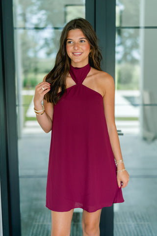 A woman wearing a sleeveless, high-neck halter-style mini dress in deep burgundy, standing in front of a modern glass door.